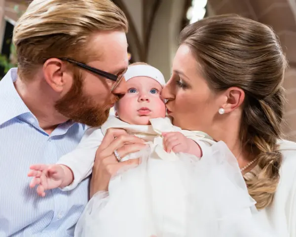 Eltern küssen ihr Baby bei der Taufe. Die Frage, wie viele Paten darf man haben, beschäftigt viele.