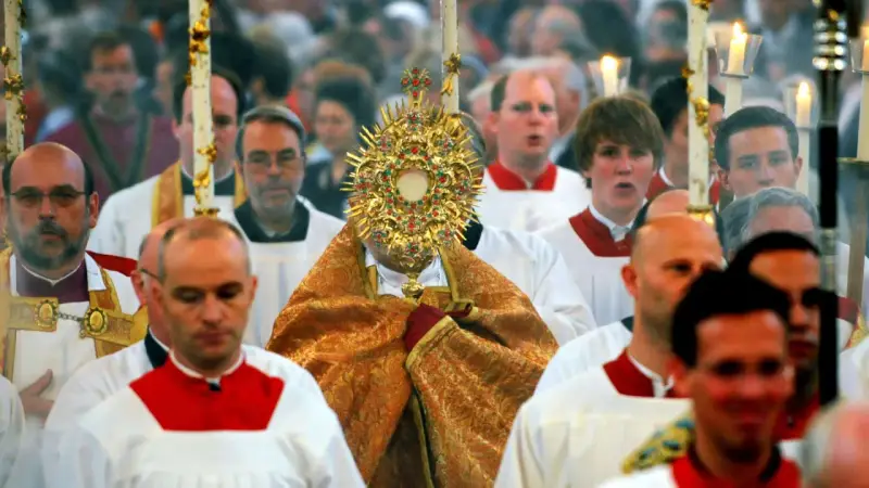 Bei Fronleichnam feiert eine Prozession mit einem goldenen Monstranz. Männer in weißen Gewändern mit roten Kragen gehen mit.